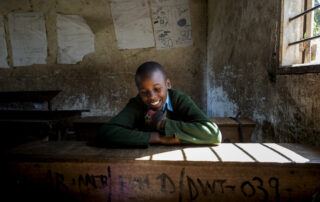 A student laughs as she sits at a wooden desk near a window at a primary school in rural Tanzania. The student is wearing a green sweater. The concrete walls are covered in paper lessons.