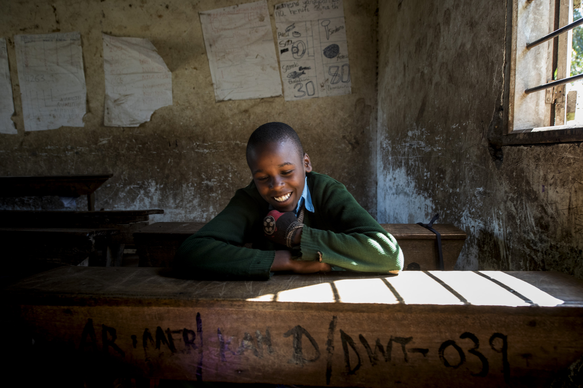 A student laughs as she sits at a wooden desk near a window at a primary school in rural Tanzania. The student is wearing a green sweater. The concrete walls are covered in paper lessons.