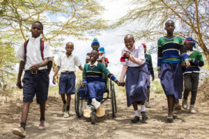 African students walk to school. One girl is in a wheelchair in the center and hold hands with another female student. They are happy and laughing.