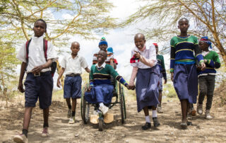 African students walk to school. One girl is in a wheelchair in the center and hold hands with another female student. They are happy and laughing.