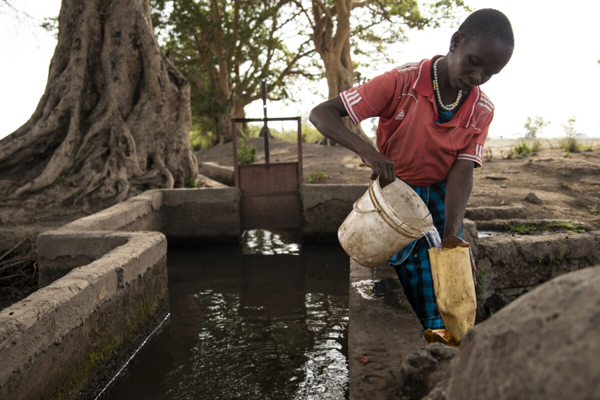 African woman collects water from a river in Tanzania. She is wearing a red shirt and holding two buckets.