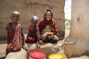 An African woman fills up her water bucket with her two sons outside her home in Tanzania. All three are dressed in red Masai shukas.