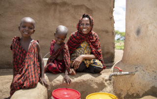 An African woman fills up her water bucket with her two sons outside her home in Tanzania. All three are dressed in red Masai shukas.
