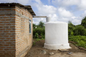 White cylindrical water tank beside a brick house in a rural yard under a cloudy sky.