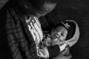 Adult cradling a newborn wrapped in a fleece blanket and wearing a knit hat, looking down at the baby in a dimly lit setting.
