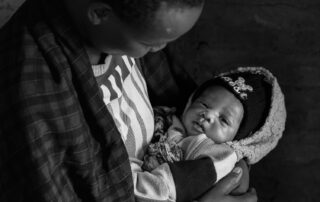 Adult cradling a newborn wrapped in a fleece blanket and wearing a knit hat, looking down at the baby in a dimly lit setting.
