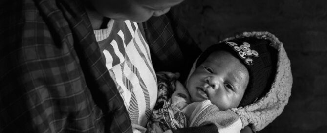Adult cradling a newborn wrapped in a fleece blanket and wearing a knit hat, looking down at the baby in a dimly lit setting.