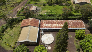 Aerial view of a rural property with rusted red metal roofs, circular water tanks, and surrounding trees and grassland.