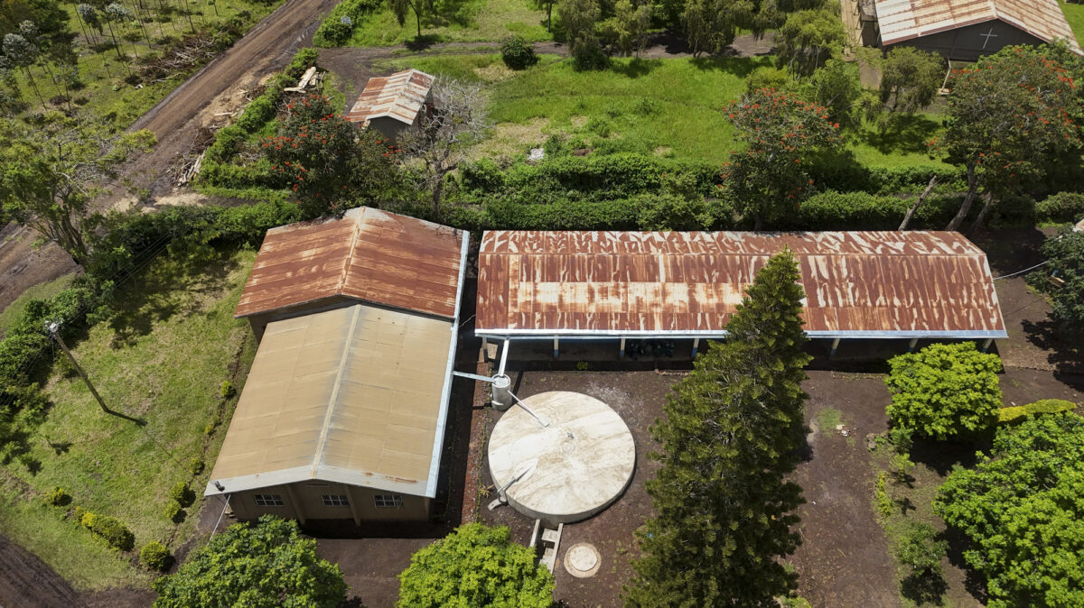 Aerial view of a rural property with rusted red metal roofs, circular water tanks, and surrounding trees and grassland.