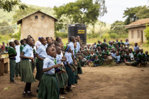 Group of schoolchildren in green uniforms standing in rows outdoors, likely during a school assembly or event in a rural area.