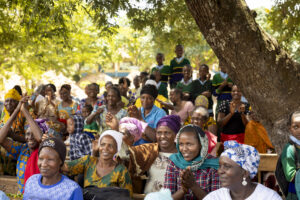 Group of people at an outdoor community gathering, smiling and clapping under a tree in bright daylight.
