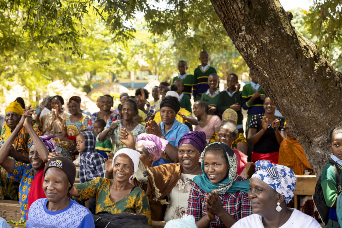 Group of people at an outdoor community gathering, smiling and clapping under a tree in bright daylight.